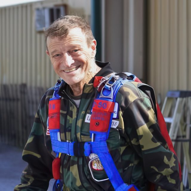 A smiling man wearing a camouflage jumpsuit stands outside, equipped with a parachute harness in bright red and blue. A small ladder and a building are visible in the background.