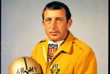 A man wearing a yellow jacket holds a football helmet with "ARMY" written on it. He has several military service ribbons on his shirt. The background is plain and light-colored.