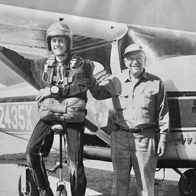 Two men smiling in front of a small aircraft. The man on the left is wearing a parachute and helmet, while the man on the right is in casual clothing and a cap. The plane's tail number is partially visible. The scene appears vintage.