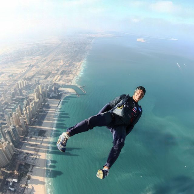 A person skydiving over a coastal city, with tall buildings casting shadows on the sandy beach below and the vast ocean stretching into the horizon.