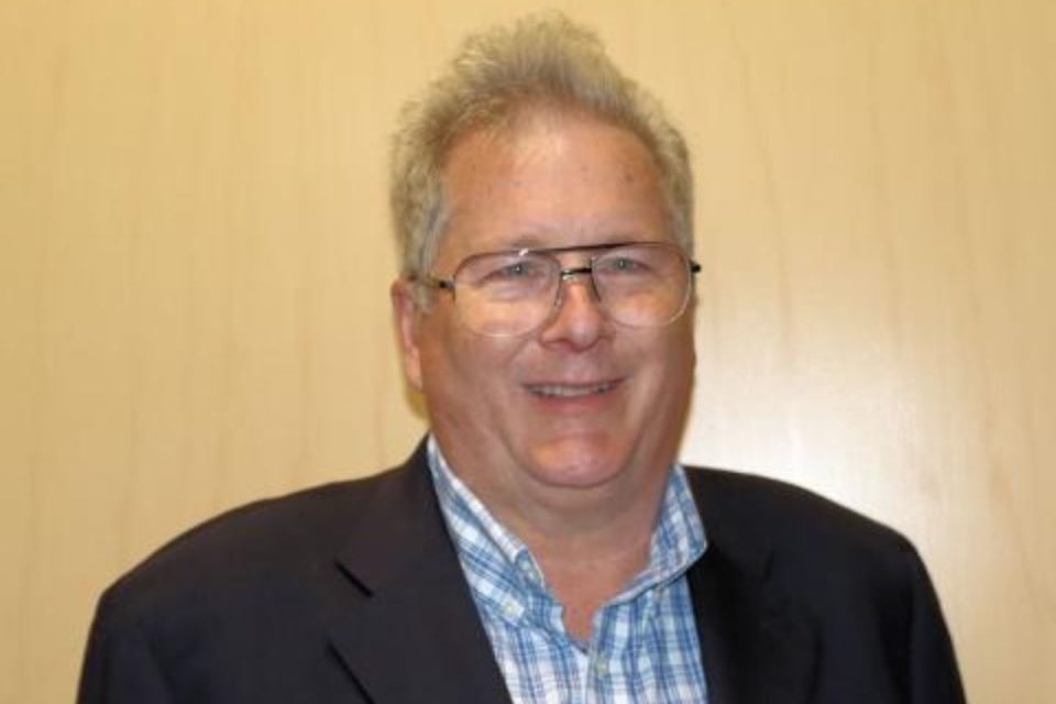 A person with short, light gray hair and glasses is smiling. They are wearing a dark jacket over a blue and white checkered shirt, standing against a plain light-colored background.