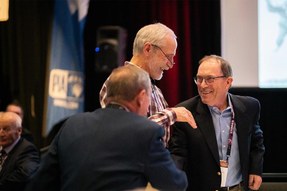 Two men greet each other warmly at an indoor event, surrounded by other attendees. They are smiling, with one extending a hand towards the other. A banner and a screen are visible in the background.