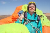 A person wearing a colorful parachuting suit and helmet smiles at the camera. They are holding the lines of a brightly colored parachute behind them, standing in an open landscape under a clear blue sky.