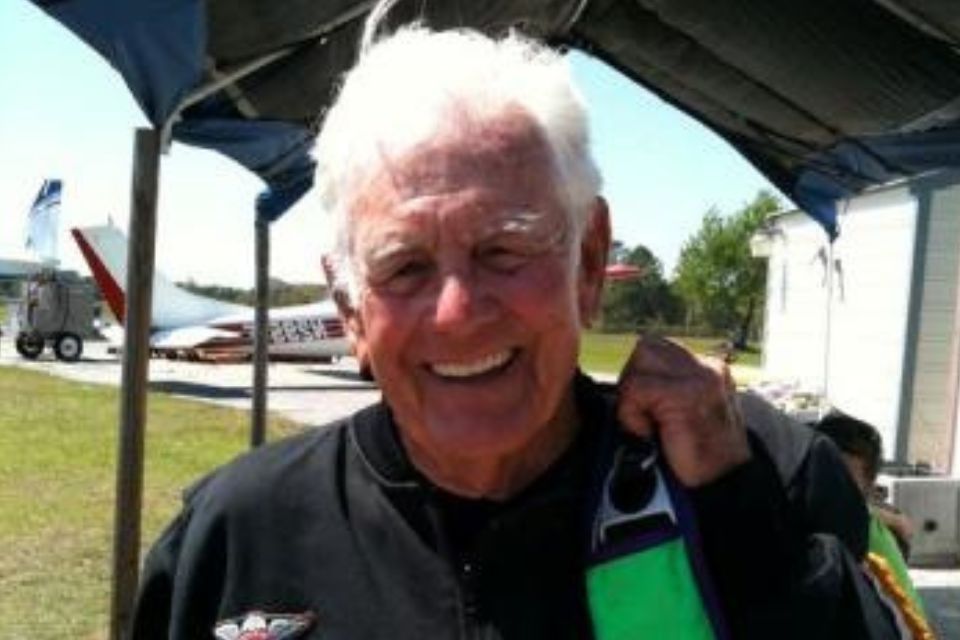 An elderly man smiling broadly, wearing a dark jumpsuit, stands under a canopy at an airfield. A small plane is visible in the background on a grassy area.