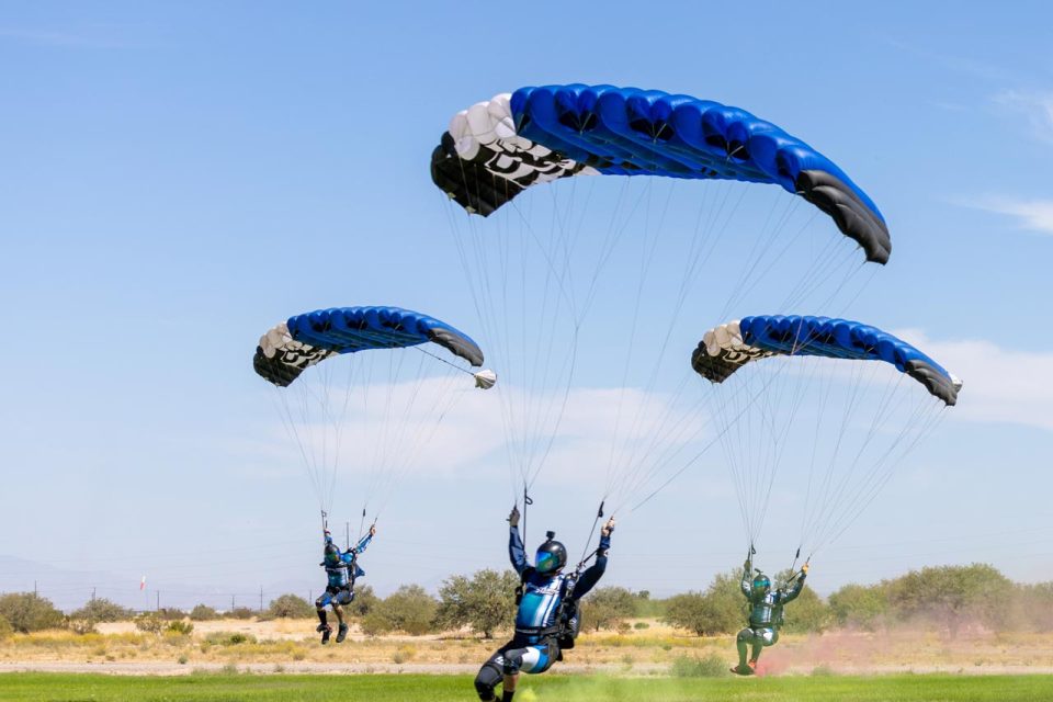 Three skydivers in blue and black gear are landing on a grassy field with their parachutes open. The sky is clear and sunny, and the landscape in the background is sparse with trees and bushes.