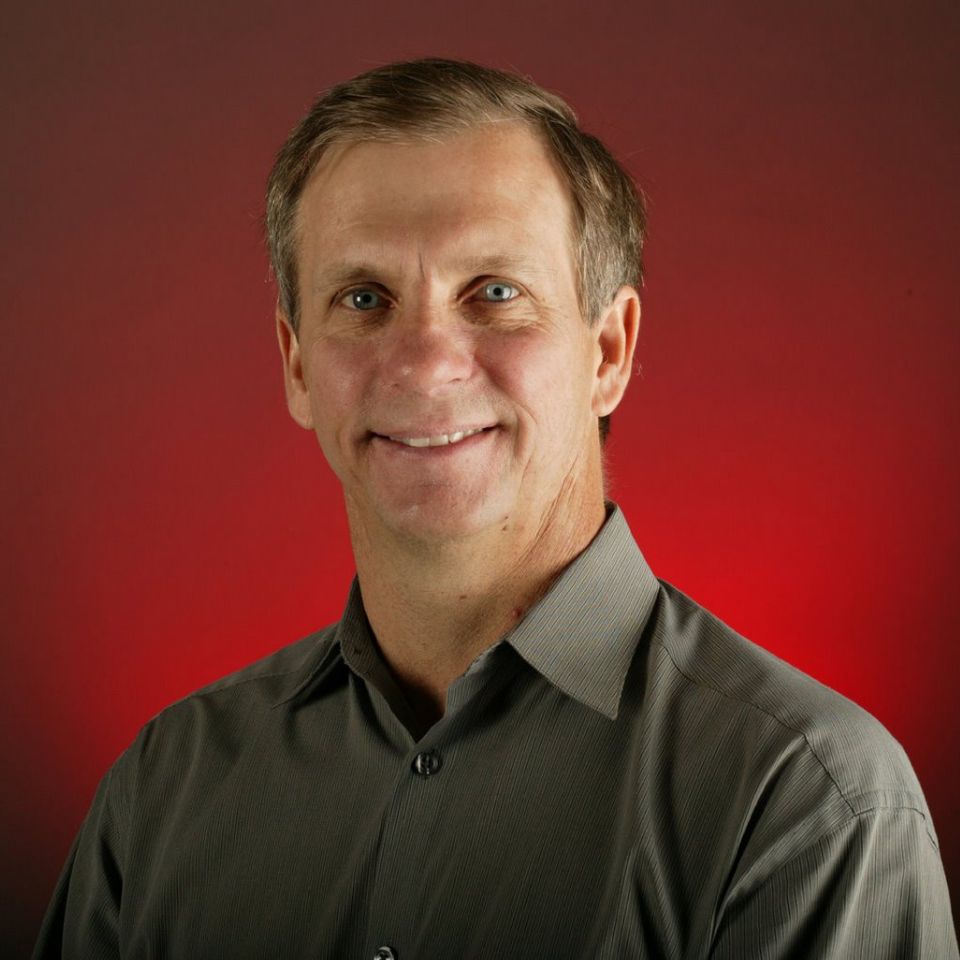 A man with short hair wearing a gray collared shirt is smiling against a red background.