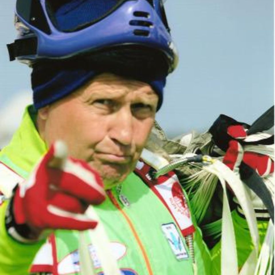 A person in a green jacket and red gloves wearing a helmet looks into the camera. They are holding parachute lines and pointing forward with a determined expression. The background is a clear blue sky.