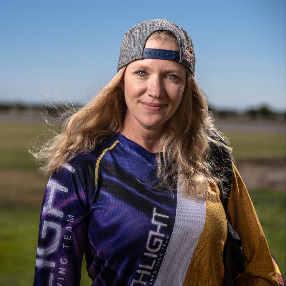 A woman with long blonde hair wears a gray cap backward and a colorful sports jersey. She stands outdoors on a sunny day, smiling confidently. The background features a clear blue sky and grassy fields.