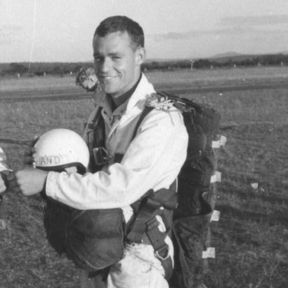 A person in a jumpsuit and harness holds a helmet and parachute pack, standing outdoors on a grassy field. The smiling person appears ready for a parachute jump, with trees and distant hills in the background.