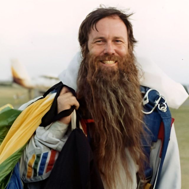 A bearded man smiling, wearing a skydiving jumpsuit with a parachute pack on his back. He is holding onto the parachute cords. A small airplane is visible in the grassy background.