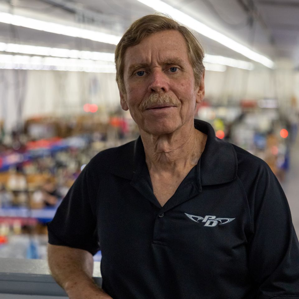 A man with a mustache, wearing a black polo shirt with a logo, stands indoors with a background of a bustling workshop or factory. He gazes directly at the camera, with overhead lights illuminating the scene.