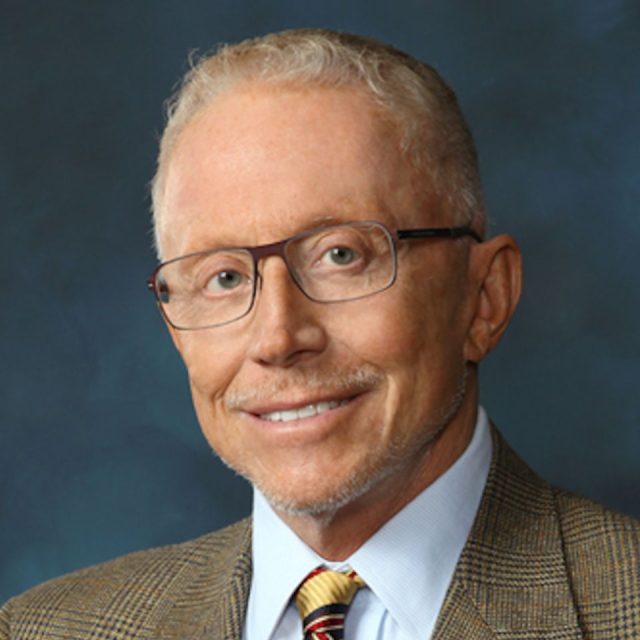 Man with short hair, glasses, and light beard, wearing a brown plaid suit jacket, blue shirt, and striped tie, smiling against a dark blue background.