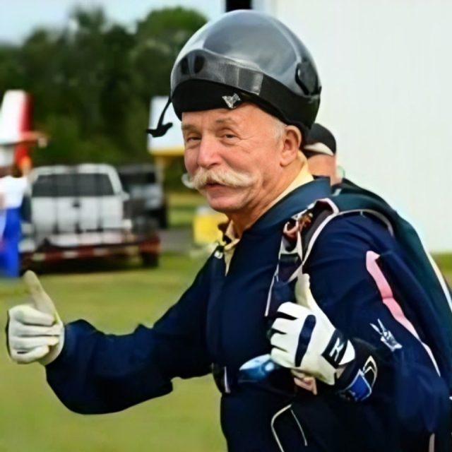 Elderly man with a large mustache wearing a skydiving suit and helmet gives two thumbs up outdoors, with vehicles and greenery in the background.