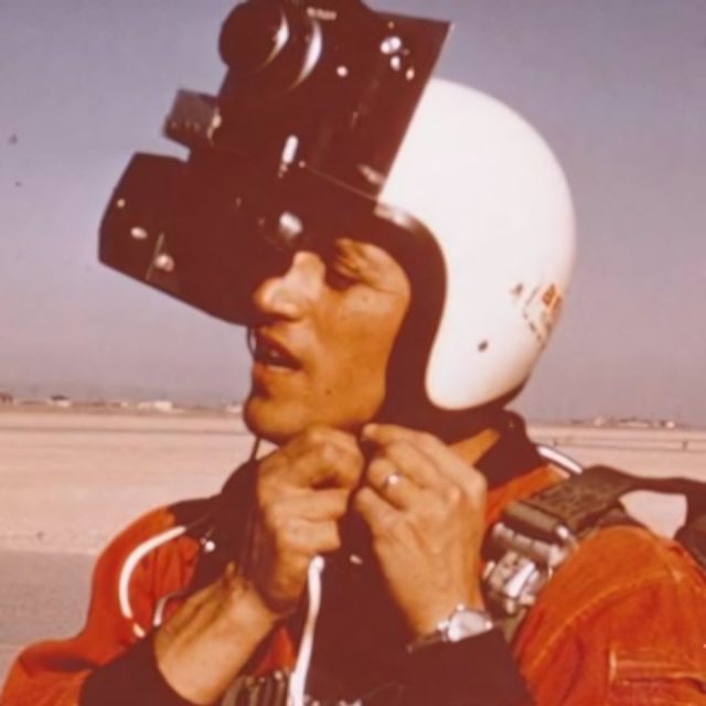 A person wearing a helmet with a large camera mounted on top adjusts their chin strap. They are outdoors, possibly at an airfield, with a vast, open landscape in the background.