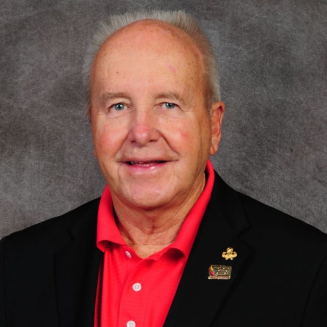 An older man with short, white hair wearing a black blazer and red polo shirt, smiling at the camera against a gray background.