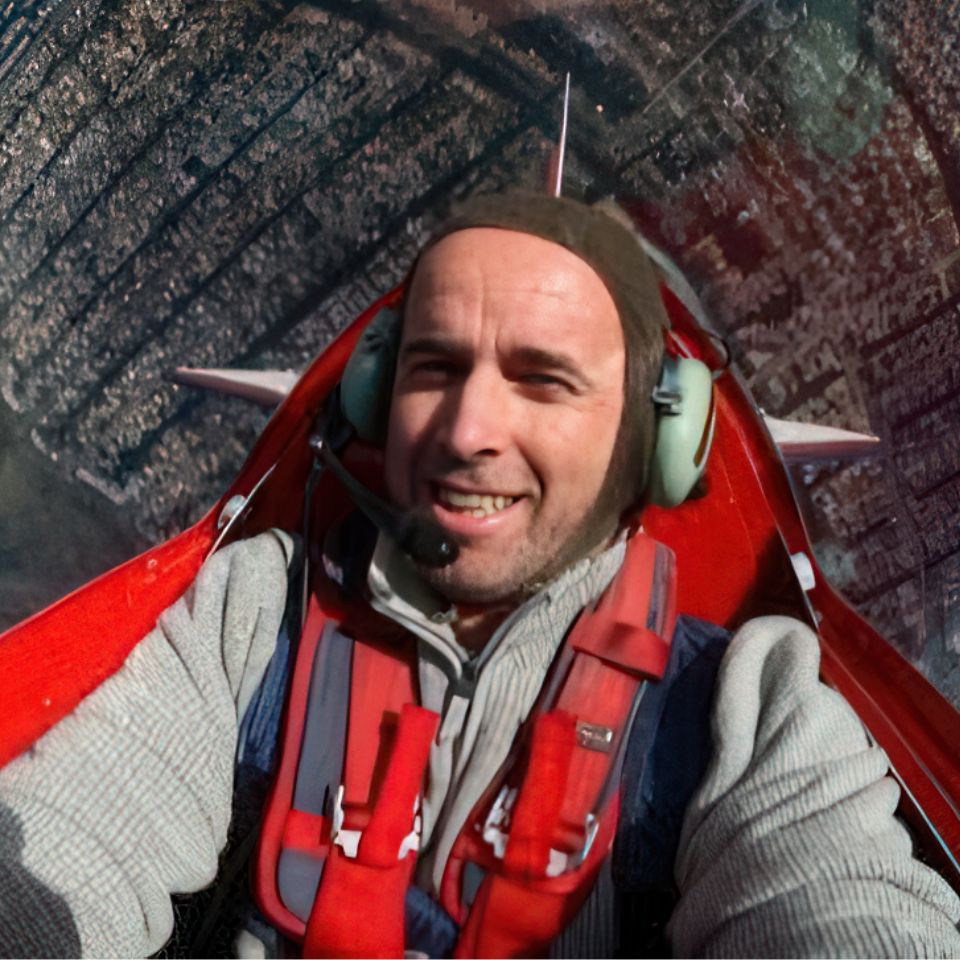 A person wearing a headset and flight gear smiles while flying in a red aircraft. The aerial view below shows a densely populated urban area.