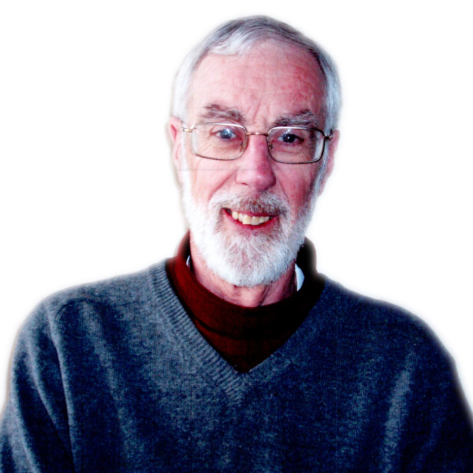 An elderly man with glasses and a white beard, wearing a gray sweater over a brown turtleneck, smiles at the camera against a plain white background.