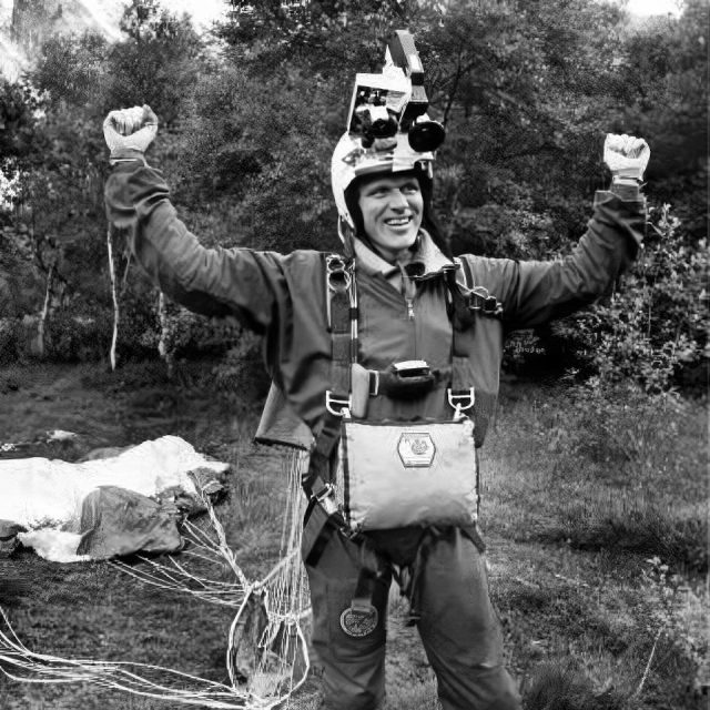 A person wearing a helmet and gear smiles with raised arms next to a parachute on the ground. They're outdoors on grassy terrain with trees in the background. The image is in black and white.