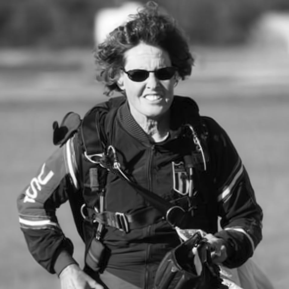 A person with short curly hair, sunglasses, and a jumpsuit prepares to skydive, holding a parachute. They appear focused and determined, with a blurred background suggesting movement or a landscape. Black and white image.