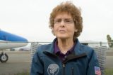 A person with curly hair and glasses sits in front of a small aircraft. They wear a blue jacket with an American flag patch and an emblem. The background shows a fence and an airfield.