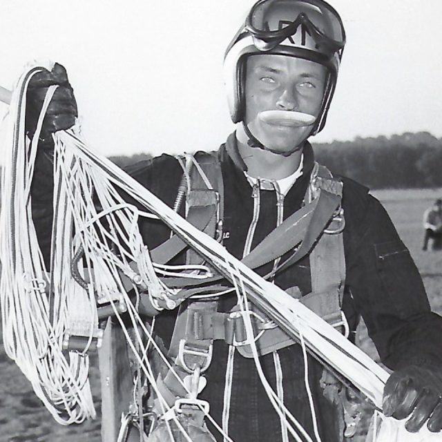 A skydiver wearing a jumpsuit and helmet stands on a field, smiling and holding open parachute lines. The background shows a blurred treeline and another person in the distance. The image is in black and white.