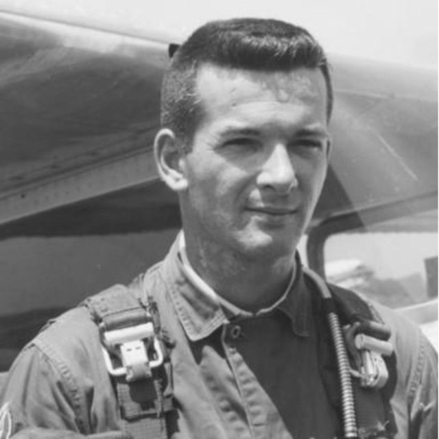 A black and white photo of a man in a flight suit standing in front of an aircraft. He has short dark hair and is wearing a parachute harness. The image has a vintage appearance.