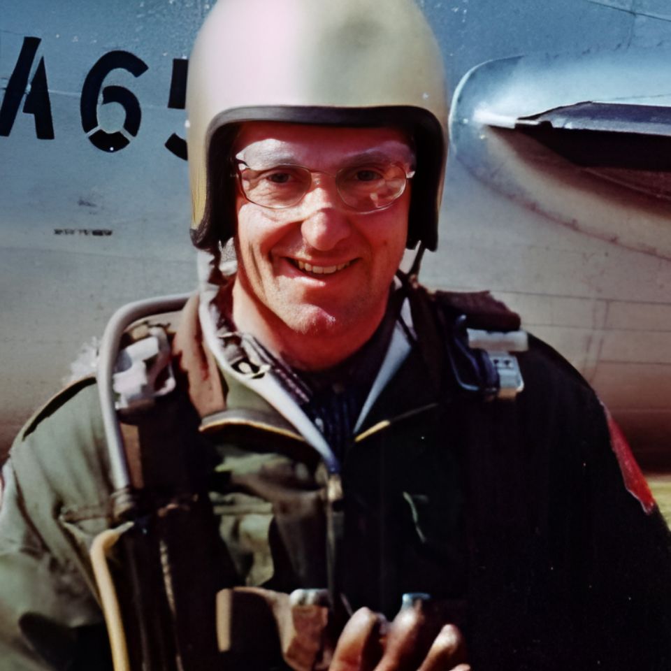 A person wearing a helmet and glasses, dressed in a green flight suit with parachute gear, stands smiling in front of an aircraft. The numbers "465" are partially visible on the plane behind them.