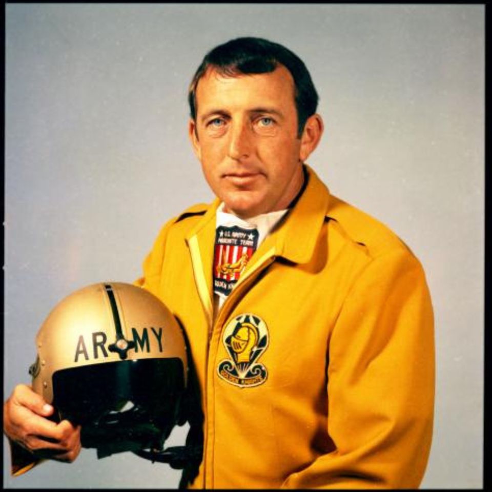 A man wearing a yellow jacket with a patch on the chest holds a helmet labeled "ARMY." He has short hair and a neutral expression, standing against a plain background.