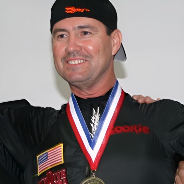 A person smiling, wearing a black shirt with patches and a black cap. They have a medal with a red, white, and blue ribbon around their neck.