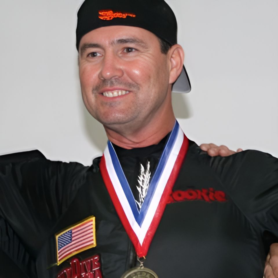 A person smiling, wearing a black shirt with patches and a black cap. They have a medal with a red, white, and blue ribbon around their neck.