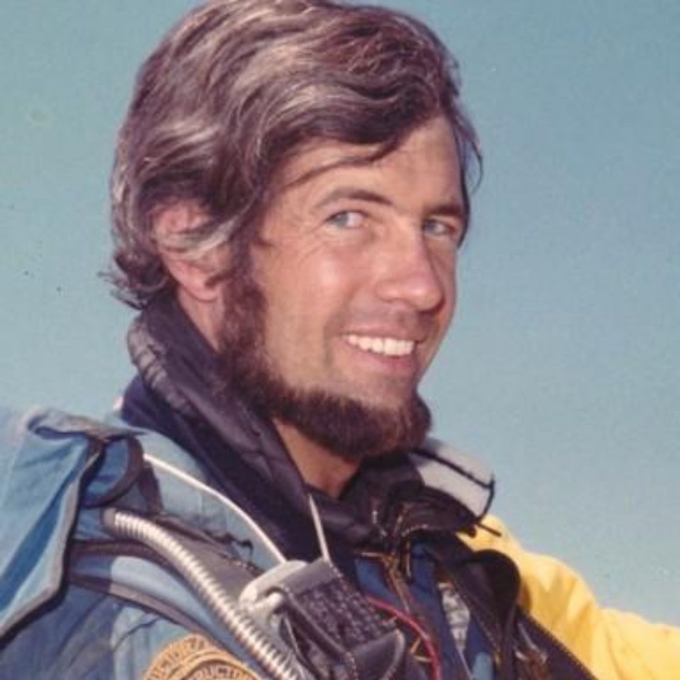 A person with a beard smiles against a clear blue sky, wearing a colorful jacket. The attire features patches and appears to be related to outdoor or adventure gear, suggesting an adventurous setting.