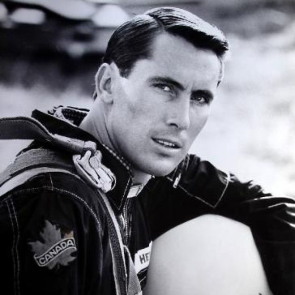 A man in a jumpsuit with a Canadian patch on his shoulder sits outdoors. The image is in black and white, highlighting his focused expression and short, styled hair.