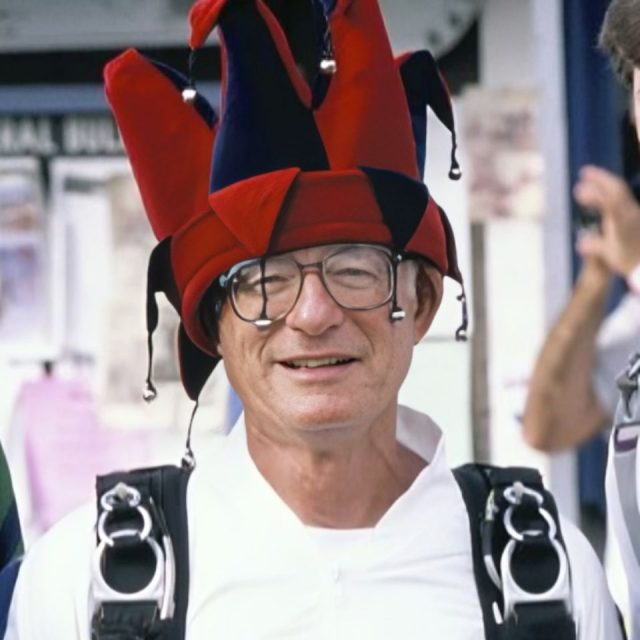 A person wearing a colorful jester hat smiles while in a skydiving harness. The background is slightly blurred.