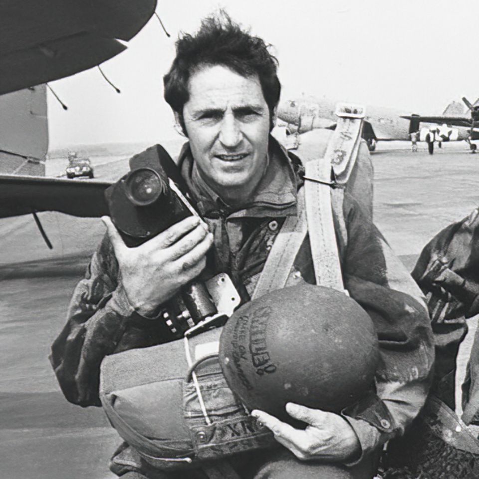 A man in military gear holds a camera and a helmet, standing on an airfield. He wears a jumpsuit with straps and buckles, and the background features aircraft. The scene has a vintage, black-and-white appearance.