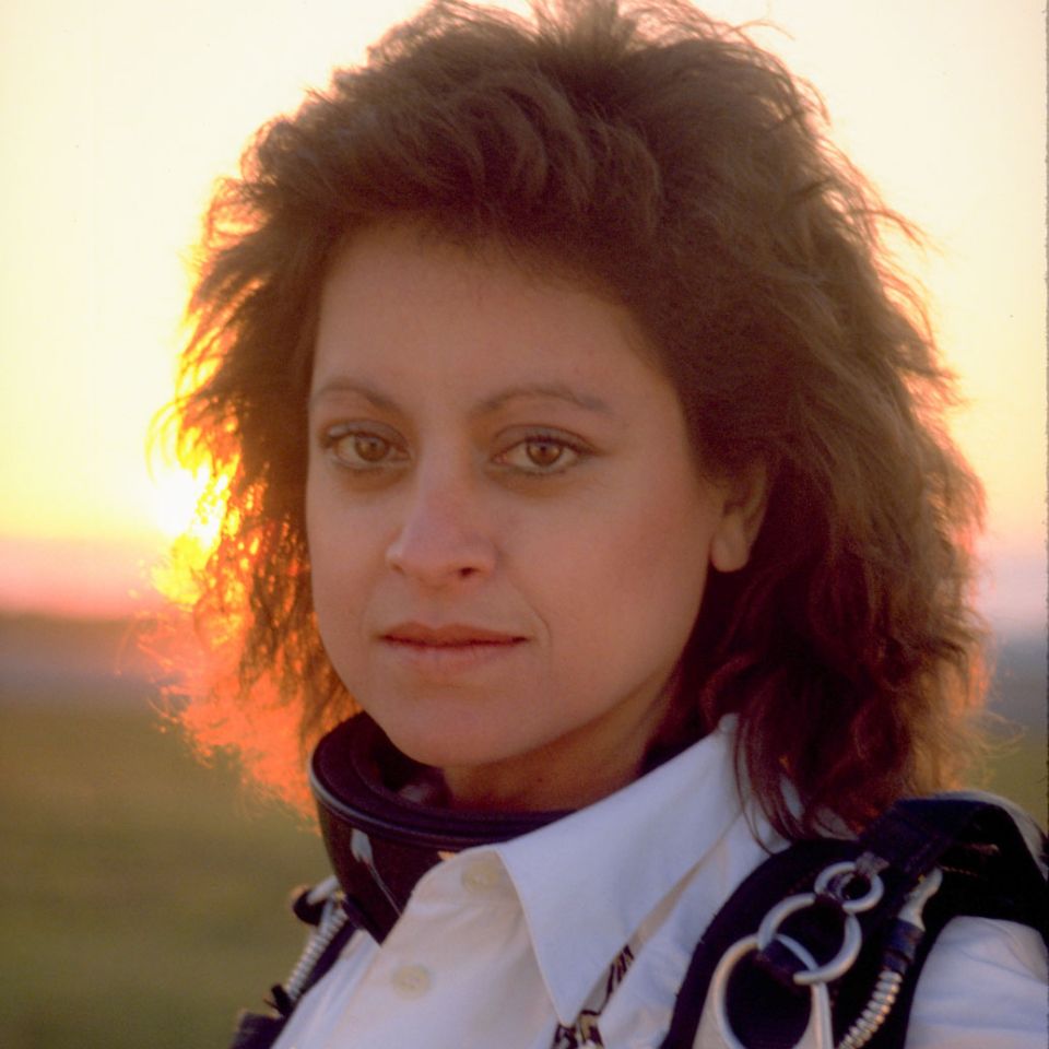 A person with curly hair stands outdoors at sunset, wearing a white shirt and parachute harness. The sun sets in the background, casting a warm glow.