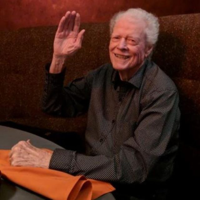 An elderly man with white hair smiles and waves while sitting at a table in a dimly lit room. He wears a dark-colored, patterned shirt, and an orange napkin is on the table in front of him.