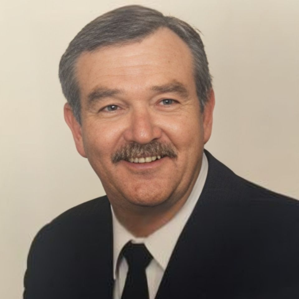 A man with short gray hair and a mustache is smiling. He is wearing a dark suit, white shirt, and dark tie against a plain light background.