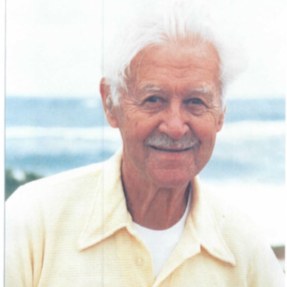 An elderly man with white hair and a mustache is smiling outdoors. He is wearing a light yellow shirt, and the background shows a glimpse of the ocean and blue sky.