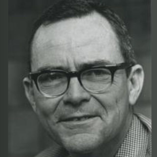 A black and white portrait of a middle-aged person wearing glasses and a checkered shirt, smiling slightly. The background is blurred.