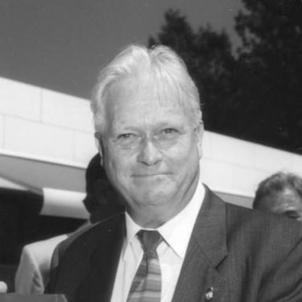 Black and white photo of a man in a suit and tie, smiling slightly. He has white hair and is standing outdoors against a backdrop of a building and trees.