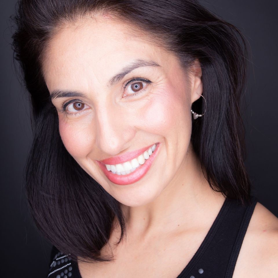 A woman with shoulder-length dark hair smiles brightly. She is wearing hoop earrings and a black top, set against a dark background. Her makeup is subtle, highlighting her natural features.