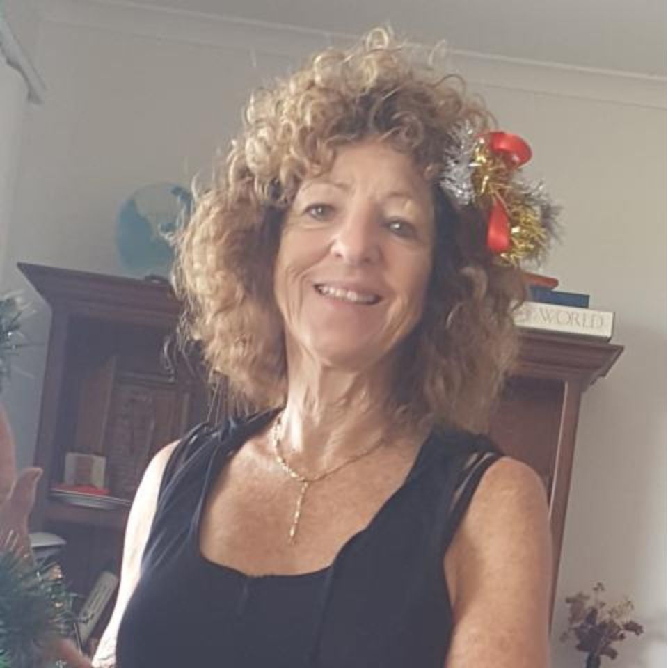 A woman with curly hair smiles at the camera, wearing a festive headband with red and gold decorations. She is in a room with a wooden cabinet in the background.