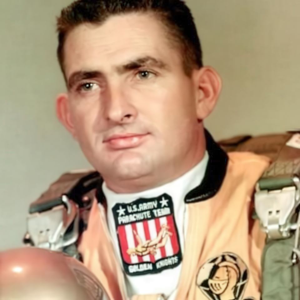 A man wearing a U.S. Army Golden Knights parachute team uniform is shown. He has short dark hair and is holding a helmet. The uniform features patches and badges related to parachuting.