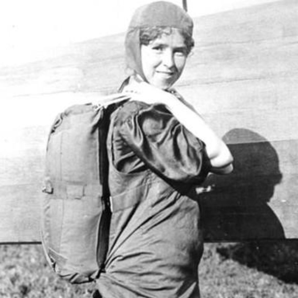 A black and white photo of a person wearing an early 20th-century aviation outfit, including a pilot cap, standing in front of an aircraft while carrying a backpack. The person is smiling and looking towards the camera.