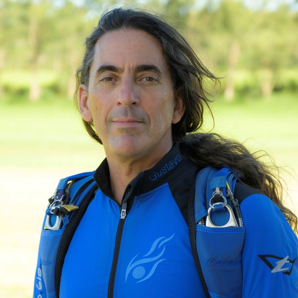 A person with long hair stands outdoors in a blue skydiving suit. They have a calm expression, and green grass and trees are blurred in the background.