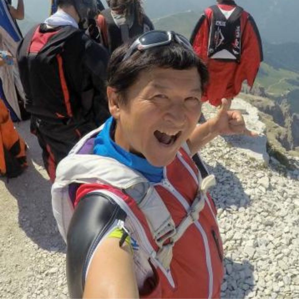 A person wearing a red wingsuit takes a selfie with a group of wingsuit flyers on a rocky mountain peak. They have a joyful expression and the background shows a scenic view of mountains and valleys under a clear sky.