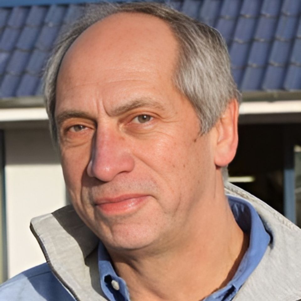 A person with short, graying hair is wearing a light blue shirt and a light-colored jacket. They are standing outdoors in front of a building with a tiled roof. The sunlight is illuminating their face.