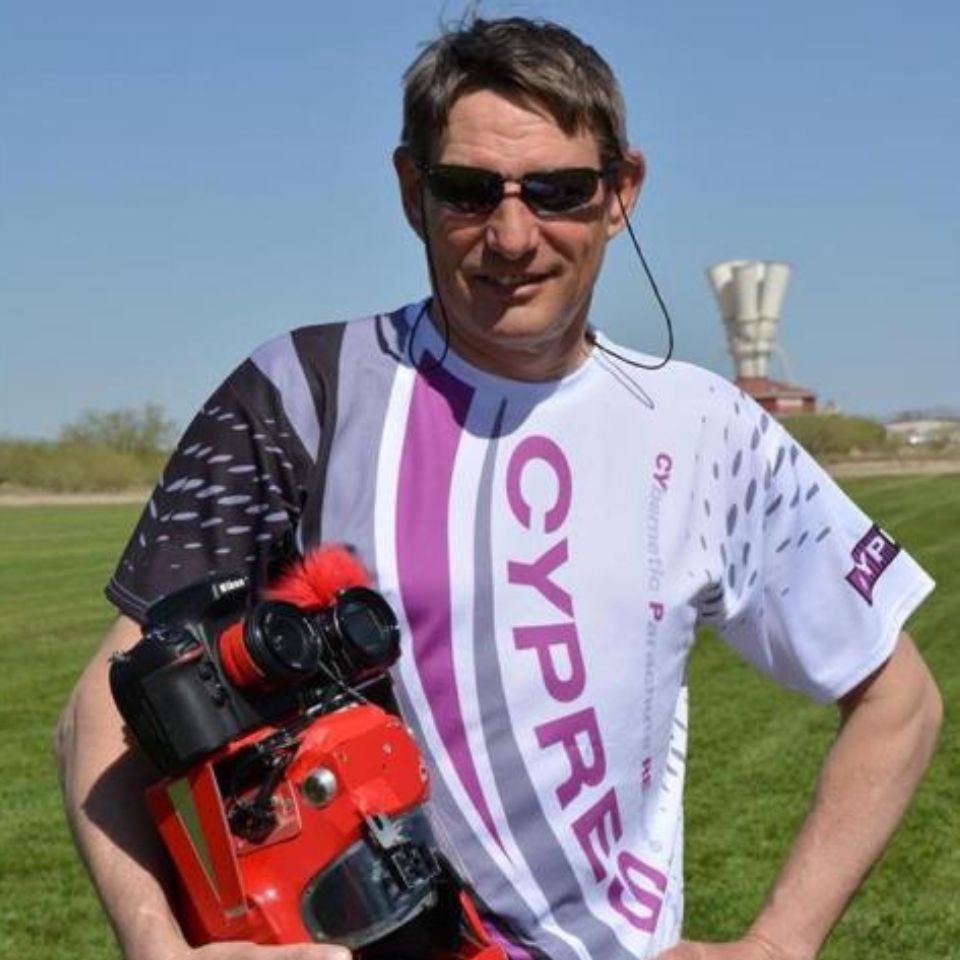 A person with sunglasses is standing outdoors on a grassy field, holding a red object with lenses. They are wearing a white and purple t-shirt with the word "CYPRES" on it.