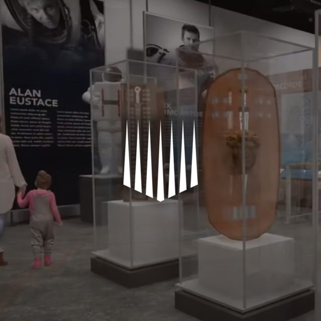 A museum exhibit showcasing various space exploration items in glass cases. A woman holds a child's hand as they view the displays, including a space suit and related artifacts. A large logo is prominently featured in the center.