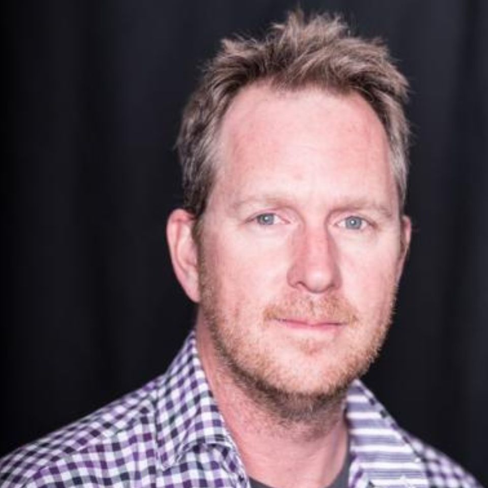 A man with short, light brown hair and a beard is wearing a checkered shirt. He stands against a dark background, looking at the camera with a neutral expression.